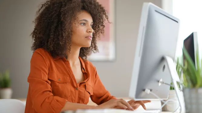 Uma pessoa com cabelos cacheados está sentada em uma mesa, trabalhando em um computador. Eles estão vestindo uma camisa laranja e aparecem focados na tela. O fundo inclui uma parede desfocada e algumas plantas.