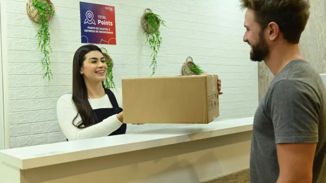 Uma recepcionista entrega um pacote de papelão a um homem sorridente do outro lado do balcão. A área da recepção tem uma parede de tijolos brancos com duas decorações de plantas penduradas e uma placa “Total de Pontos”. Ambos os indivíduos parecem estar em uma interação alegre.