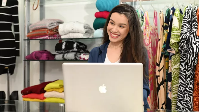 Uma mulher está sorrindo enquanto está sentada diante de um laptop em uma loja de roupas. As prateleiras atrás de sua vitrine dobravam roupas e várias peças penduradas.