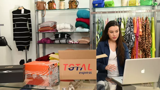 Uma mulher está sentada em uma mesa usando um laptop em uma loja de roupas. Há roupas coloridas nas prateleiras ao fundo. Vários pacotes, incluindo uma caixa chamada “Total Express”, estão sobre a mesa, sugerindo que ela está preparando itens para envio.