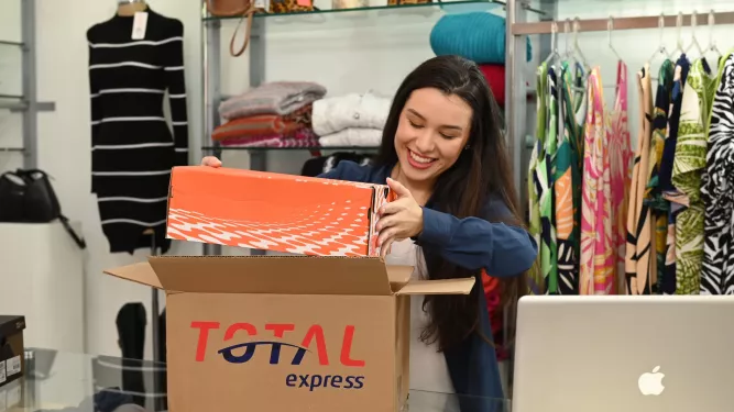 Uma mulher com longos cabelos castanhos sorri enquanto abre uma caixa de sapatos laranja em uma loja de roupas. Ela está atrás de um balcão com um laptop e cercada por diversas roupas e acessórios. Uma caixa de remessa Total Express aberta está na frente dela.