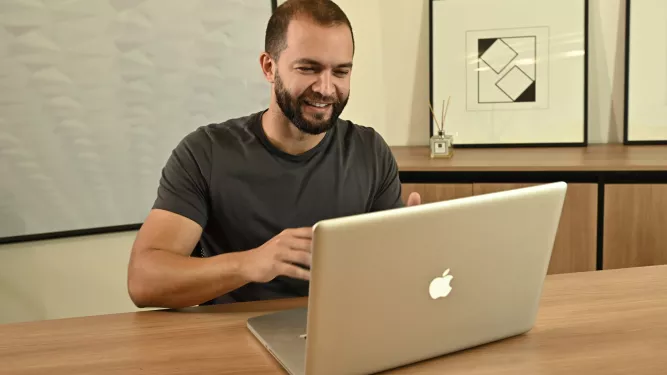 Um homem com barba sorri enquanto usa um laptop em uma mesa. Há estampas geométricas emolduradas e um difusor de palheta no fundo.
