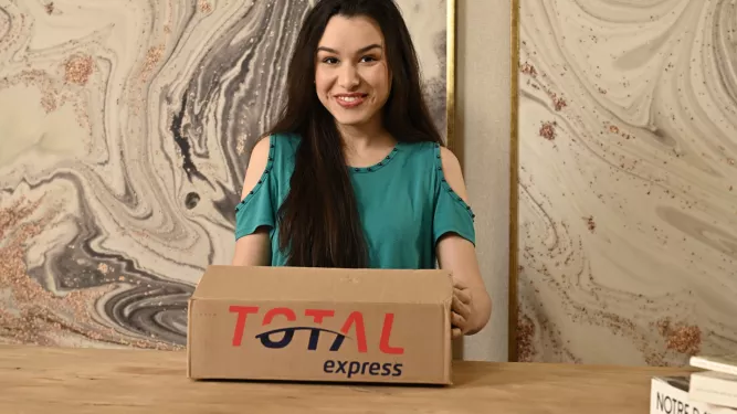 Uma mulher com longos cabelos escuros está sorrindo e segurando uma caixa de papelão com a inscrição "TOTAL express" enquanto está sentada em uma mesa de madeira. O fundo apresenta um desenho de parede em mármore. Um livro com o título "NOTRE DAME" está parcialmente visível na mesa à direita.