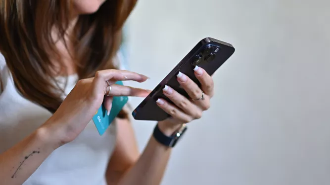 Close de uma mulher usando um smartphone. Ela está vestindo uma camisa branca e vários anéis e pulseiras. Ela está segurando um cartão de crédito na mão esquerda, com a mão direita tocando a tela do telefone. Seu rosto está parcialmente visível.