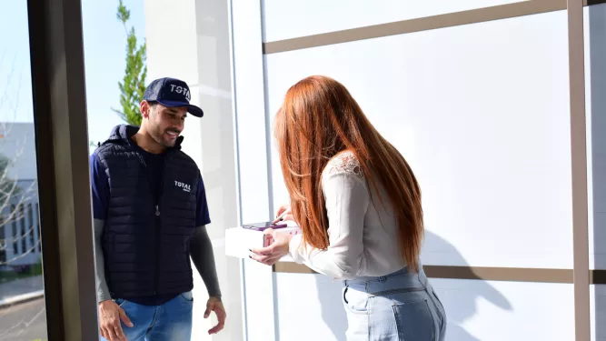 Um entregador de boné e colete escuros entrega um pacote a uma mulher de longos cabelos ruivos. A mulher, vestida com blusa clara e calça jeans, usa uma caneta para assinar a entrega em um dispositivo portátil. Eles estão em uma sala bem iluminada com grandes janelas.