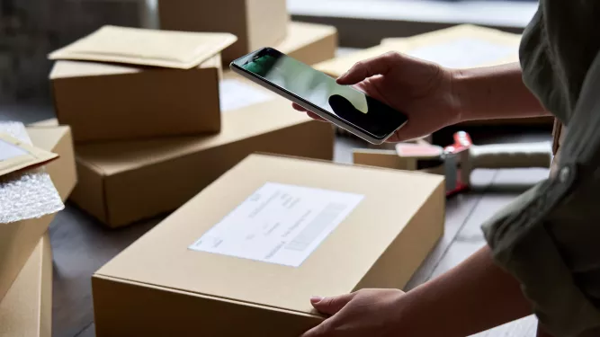 Pessoa usando um smartphone para tirar uma foto de uma caixa marrom embalada com uma etiqueta de remessa. Várias outras caixas e envelopes estão espalhados sobre a mesa. Um dispensador de fita também é visível ao fundo.