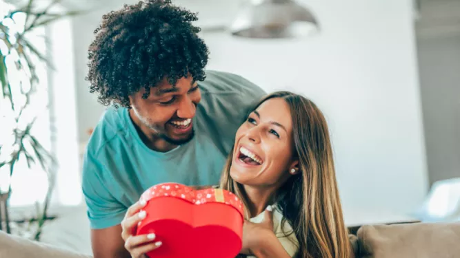 Um casal sorri alegremente enquanto segura uma caixa de presente vermelha em formato de coração. O homem está atrás de uma mulher que segura a caixa, e eles parecem estar em uma sala moderna e iluminada. Uma planta e uma luminária são visíveis ao fundo.