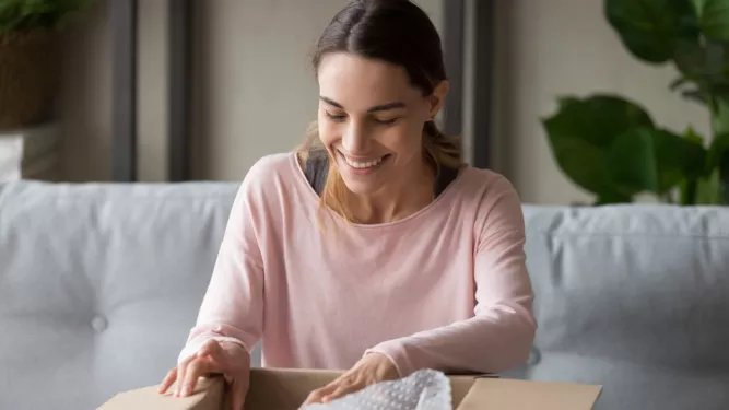 Uma mulher sorridente, vestindo uma camisa rosa de mangas compridas, está sentada em um sofá e abrindo uma caixa de papelão. Ela parece feliz e animada ao olhar dentro da caixa, com plástico-bolha parcialmente visível dentro. Uma planta verde está ao fundo.