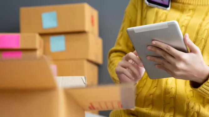 Uma pessoa vestindo um suéter amarelo usa um tablet enquanto está sentada entre várias caixas de papelão, possivelmente envolvida em um negócio on-line ou gerenciamento de estoque. As caixas possuem rótulos coloridos e uma caixa em primeiro plano está aberta.