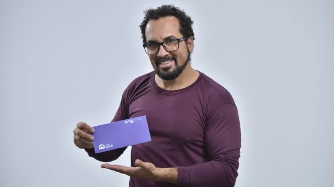 Um homem de cabelos cacheados, barba e óculos, vestindo uma camisa marrom de mangas compridas, segura e aponta para um envelope roxo com um sorriso. O fundo é totalmente branco.