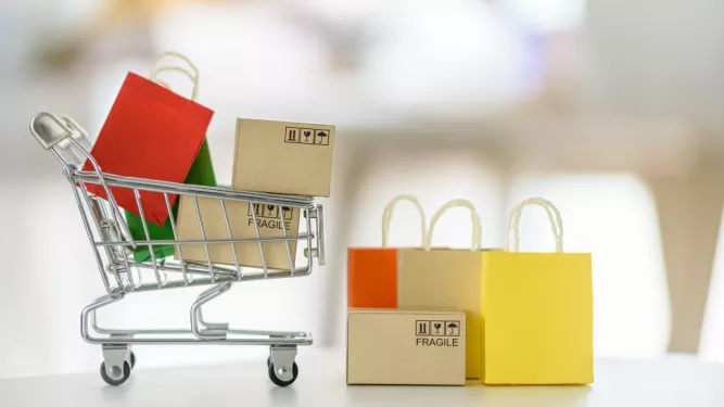 Um pequeno carrinho de compras cheio de caixas de papelão e sacolas coloridas fica sobre uma superfície branca. Ao lado, mais caixas de papelão com a etiqueta “FRÁGIL” são cercadas por sacolas de compras vermelhas, amarelas e laranja, representando um conceito de entrega de compras online.