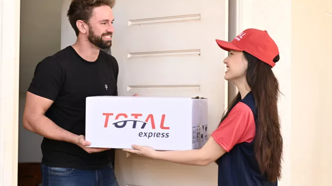 Um entregador de boné vermelho e uniforme azul entrega um grande pacote branco com o rótulo "TOTAL express" para um homem sorridente de camisa preta em sua porta.