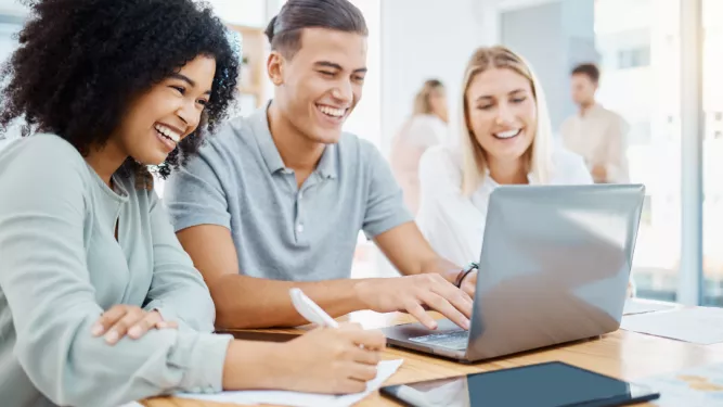 Três pessoas sorridentes sentadas ao redor de uma mesa com um laptop, colaborando e fazendo anotações. Eles parecem estar em um ambiente de escritório moderno e bem iluminado, com grandes janelas ao fundo. Papéis e um tablet também ficam visíveis sobre a mesa.
