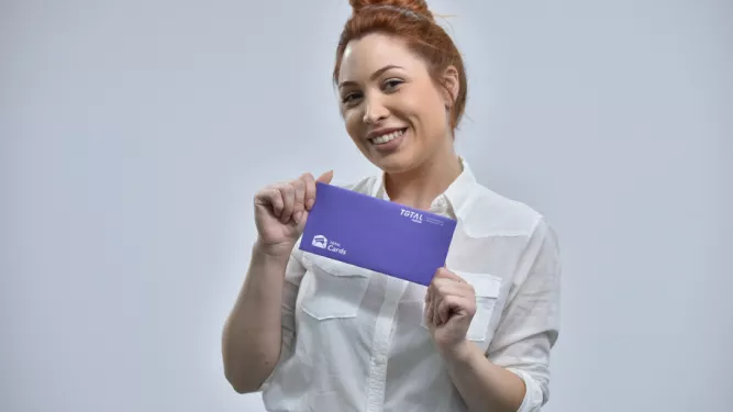 Uma pessoa com penteado em coque está sorrindo e segurando um envelope roxo com as duas mãos. Eles estão vestindo uma camisa branca de botões e contra um fundo cinza claro. O envelope contém texto e logotipos em branco.
