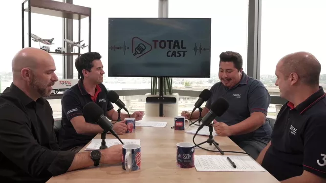 Quatro homens estão sentados ao redor de uma mesa com microfones, conversando, gravando um podcast intitulado “Total Cast”. O fundo apresenta uma grande janela com vista externa, uma tela com a logomarca do podcast e uma estante com itens decorativos.