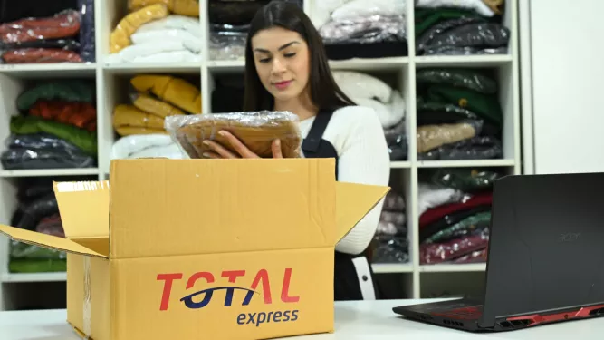 Uma mulher está atrás de um balcão, examinando uma peça de roupa embalada ao lado de uma caixa aberta do Total Express. Ao fundo, prateleiras com roupas dobradas. Um laptop está aberto no balcão.