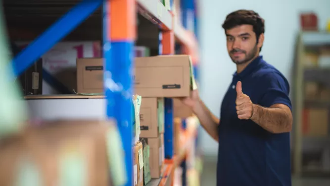 Um homem de camisa pólo azul está em um armazém, organizando caixas em uma prateleira. Ele faz um sinal de positivo com a mão direita enquanto olha para a câmera, indicando satisfação ou aprovação. Vários rótulos e embalagens coloridas são visíveis ao seu redor.