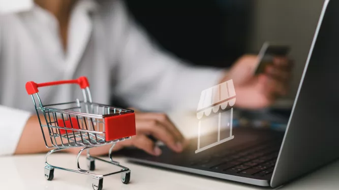 Uma pessoa segurando um cartão de crédito com uma das mãos e usando a outra para digitar no teclado de um laptop. Um pequeno carrinho de compras de brinquedos é colocado na mesa próxima. Um ícone de uma loja aparece na tela do laptop, representando compras online.