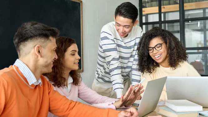 Um grupo de quatro colegas diversos está reunido em torno de uma mesa, envolvidos em uma discussão animada. Duas mulheres e dois homens estão sorrindo e interagindo entre si, com uma mulher usando um laptop. O cenário parece ser um escritório casual ou uma sala de reuniões.
