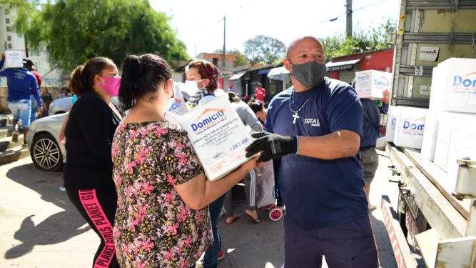 Várias pessoas usando máscaras distribuem e recebem caixas de mantimentos de um caminhão em uma rua. Um homem de camisa azul e luvas entrega uma caixa com a inscrição "Domilici" para uma mulher de camisa floral. Outros indivíduos ficam próximos com caixas.