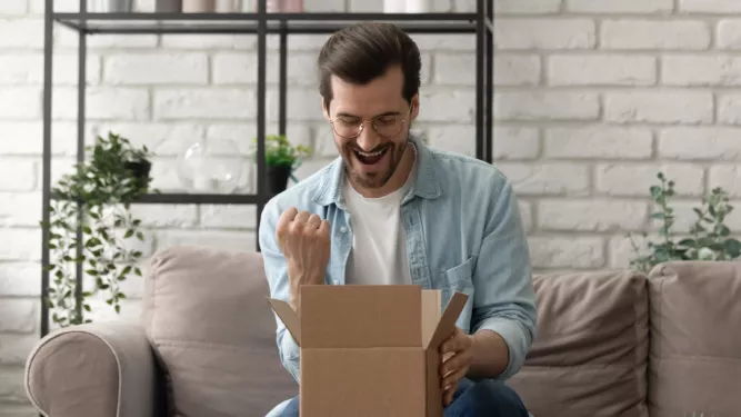 Um homem de óculos e barba, vestindo uma camisa azul clara sobre uma camiseta branca, está sentado em um sofá em frente a uma caixa de papelão aberta, parecendo animado e cerrando o punho em triunfo. Prateleiras com plantas e decoração ficam ao fundo.