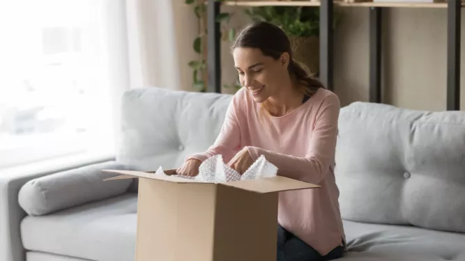 Uma mulher com longos cabelos castanhos está sentada em um sofá cinza e sorrindo enquanto abre uma caixa de papelão. A sala tem luz entrando por uma janela à esquerda e estantes com livros e plantas ao fundo. Ela parece encantada com o conteúdo da caixa.