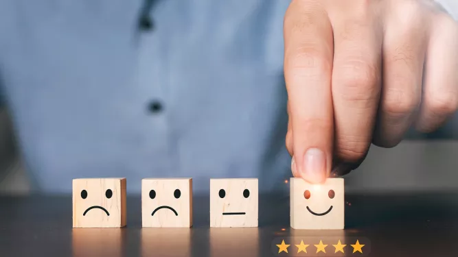 Uma pessoa organiza quatro blocos de madeira, cada um exibindo emoticons diferentes: triste, neutro, levemente sorridente e muito feliz. A pessoa toca o bloco muito feliz, com cinco estrelas douradas abaixo, significando excelente avaliação ou satisfação.