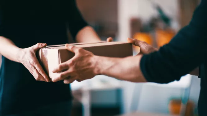 Close de duas pessoas trocando uma caixa de papelão dentro de casa. A pessoa da esquerda está entregando a caixa para a pessoa da direita. O fundo está desfocado, focando a atenção na troca de encomendas.