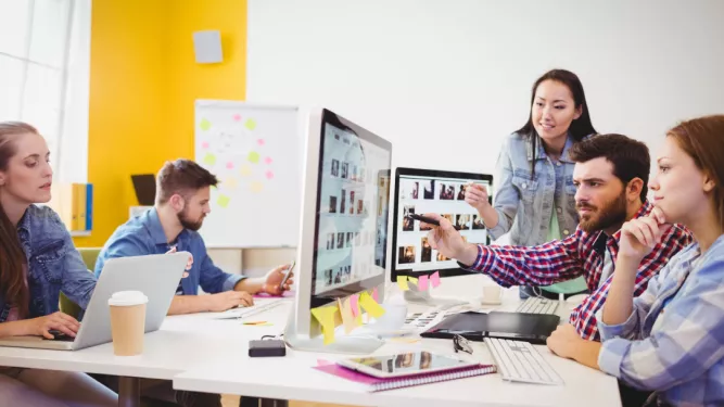 Um grupo de cinco pessoas trabalhando em um escritório moderno. Três indivíduos estão reunidos em torno de duas telas de computador, discutindo algo atentamente. Outros dois estão trabalhando em laptops em uma mesa próxima. A sala tem paredes amarelas e brancas brilhantes e uma janela com luz natural.