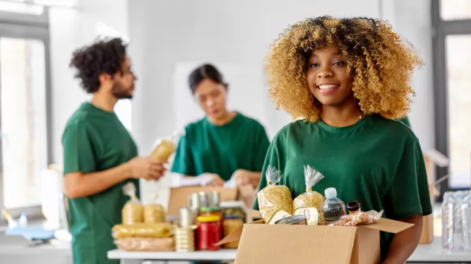 Uma mulher sorridente com cabelos cacheados segura uma caixa cheia de vários alimentos, incluindo massas e enlatados. Ao fundo, outras duas pessoas vestindo camisas verdes separam itens semelhantes sobre uma mesa. Eles parecem estar trabalhando como voluntários em um banco de alimentos.