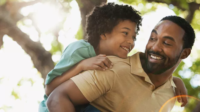 Filho e pai pretos abraçados e sorrindo, a criança está com verde e o pai com camiseta marrom, o fundo contém uma árvore. 
