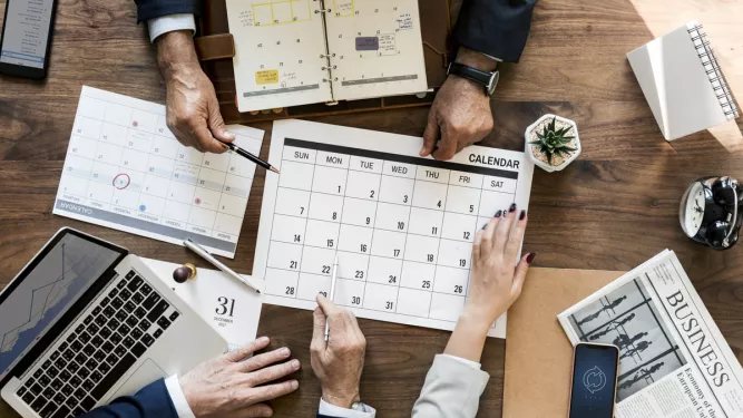 Na imagem vemos várias mãos em cima de uma mesa mexendo no calendário comercial.