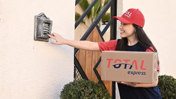 Uma pessoa tocando a campainha de uma casa para realizar uma entrega. Nas mãos, vemos uma caixa com a logo da Total Express.
