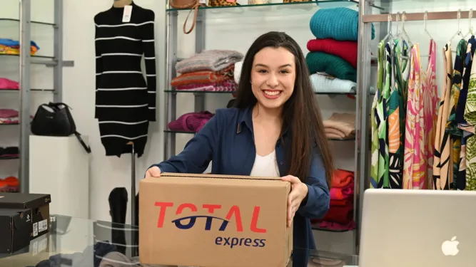 Jovem empreendedora sorridente segura uma caixa de entrega em sua loja de moda, ilustrando a etapa de envio e a praticidade de como vender roupas na internet.
