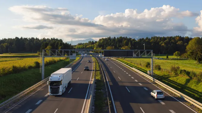 Um caminhão branco de carga viaja em uma rodovia de várias faixas no final da tarde, passando por campos e florestas com montanhas distantes, exemplificando a infraestrutura e a logística do transporte doméstico.