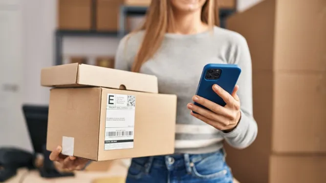 Mulher sorridente segura caixas de encomendas e um smartphone, representando a facilidade de exercer o direito de arrependimento em compras online.