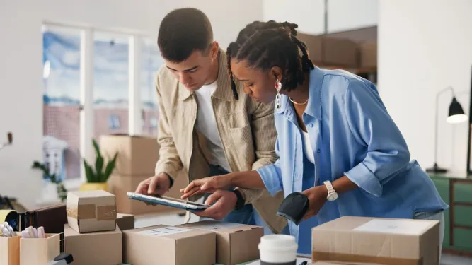A gestão de pedidos no e-commerce é ilustrada por um casal de jovens empreendedores conferindo caixas de encomenda com um tablet e um leitor de código de barras em um escritório.