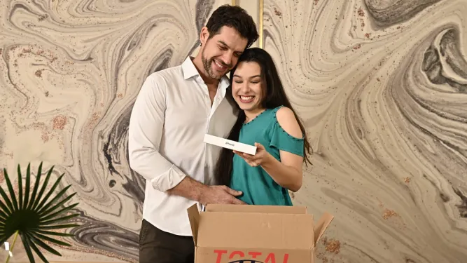 Casal sorridente abraçado abre uma caixa de encomendas da Total Express e descobre um iPhone, ilustrando a eficiência da entrega no Dia dos Namorados.