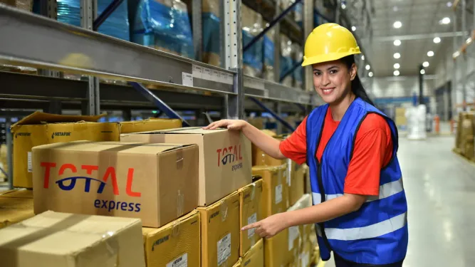 Funcionária com capacete e colete de proteção organiza caixas da Total Express em um galpão de estoque, ilustrando a operação de fulfillment ou logística própria.