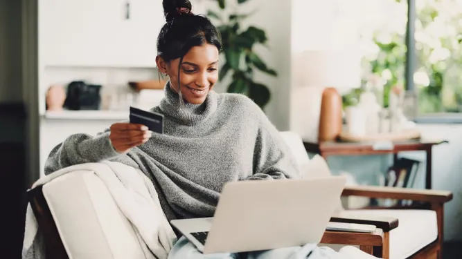 Mulher sorridente sentada no sofá usa um notebook e segura um cartão de crédito, ilustrando as novas tendências em meios de pagamento.