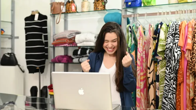 Mulher celebra no notebook em loja de roupas ao usar estratégias para vender mais.
