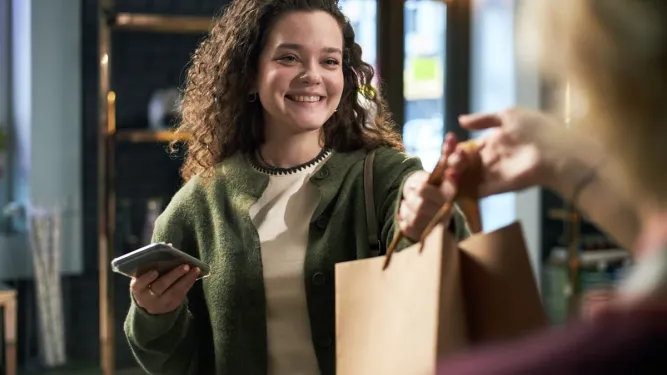 Mulher sorridente com celular recebe sacola de compras em loja no Dia do Cliente.