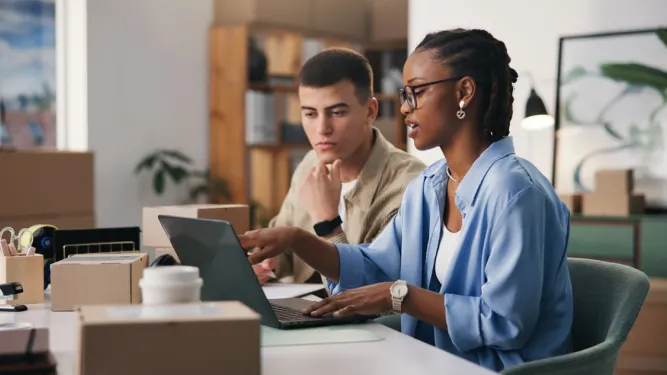 Duas pessoas analisando dados no notebook entre caixas sobre obrigação fiscal no e-commerce.