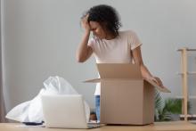 Uma mulher está ao lado de uma mesa, segurando a cabeça em frustração enquanto olha para uma caixa de papelão aberta. Ao lado dela está um laptop e alguns materiais de embalagem, sugerindo que ela está desempacotando ou embalando itens. O fundo possui uma estante parcialmente montada.
