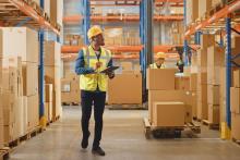 Operador de logística com colete e capacete usa tablet para conferir caixas em um galpão, realizando um inventário cíclico de mercadorias.