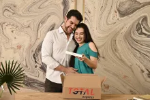 Casal sorridente abraçado abre uma caixa de encomendas da Total Express e descobre um iPhone, ilustrando a eficiência da entrega no Dia dos Namorados.