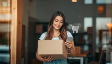 Mulher sorridente segura uma caixa de encomenda e um smartphone com ícone de carrinho, representando uma excelente experiência de compra.