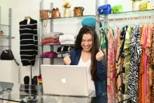 Mulher celebra no notebook em loja de roupas ao usar estratégias para vender mais.