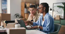 Duas pessoas analisando dados no notebook entre caixas sobre obrigação fiscal no e-commerce.