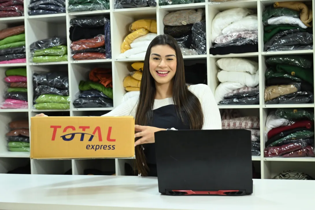 Uma pessoa com cabelos longos e escuros, vestindo blusa branca e avental escuro, segura uma caixa Total Express em frente a um laptop. Atrás deles, prateleiras estão cheias de roupas cuidadosamente dobradas de várias cores.